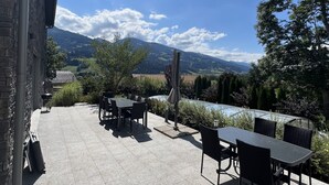 Een terras buiten met stenen vloer, tafels en stoelen, met uitzicht op een schilderachtig landschap met bergen en een waterpartij.