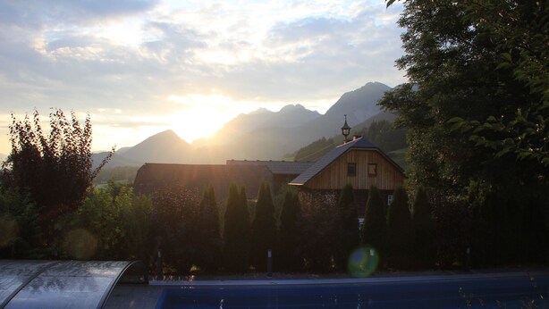 Ein rustikales Gebäude mit schrägem Dach, umgeben von Bäumen und Bergen im Hintergrund.