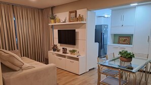 Ein modernes Wohnzimmer mit einer Couch, einem an einer weißen Kommode befestigten Fernseher, einem Esstisch mit Stühlen sowie einer Küchenzeile mit Schränken und einem Kühlschrank.
