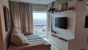 Ein modernes Wohnzimmer mit einem beigefarbenen Sofa, einem Flachbildfernseher auf einer weißen Unterhaltungseinheit und einem großen Fenster mit Vorhängen.