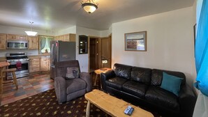 Een ruimtelijke zithoek met een kitchenette, een grijze fauteuil, een zwarte leren bank en een houten salontafel.