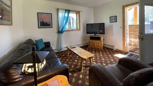 Een woonkamer met een bank, een salontafel, een televisie op een houten meubel, een raam met gordijnen en een deur naar buiten.