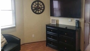 Ein Zimmer mit einem großen Flachbildfernseher an der Wand, einer schwarzen Kommode mit Schubladen, einer dekorativen Uhr und einem Fenster mit Jalousien.