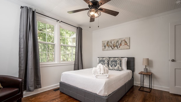 A bedroom with a bed, a ceiling fan, a window with curtains, a painting, a bedside table, and a lamp. - Enjoy the Fall & Winter Months in This Charming Home in Beautiful Marietta  (Marietta)