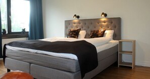 A modern bedroom with a tufted headboard, a grey bed with white linens, two black lamps on the headboard, and a small wooden nightstand beside the bed. - Millners Plaza (Tibro)