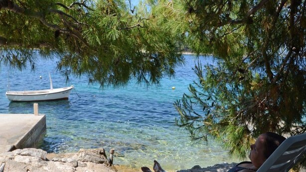 En person som kopplar av i en strandstol vid vattnet, med en båt i bakgrunden och träd ovanför.