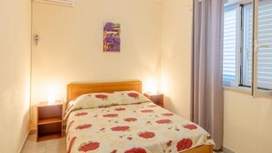 Une chambre à coucher comprenant un lit, des tables de chevet, un climatiseur fixé au mur, une fenêtre avec des rideaux et un petit élément de décoration murale.
