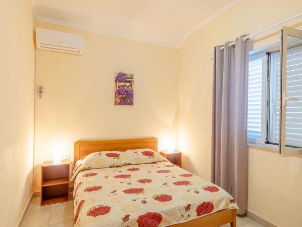 Un dormitorio con cama, mesitas de noche, aire acondicionado instalado en la pared, ventana con cortinas y una pequeña decoración en la pared.