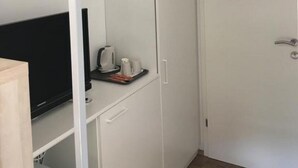 Una habitación compacta con un armario blanco, una mesita con tetera y tazas, y un televisor en un estante.