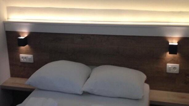 Habitación de hotel con cama, dos almohadas, una toalla doblada con cuidado y mesitas de noche fijadas a la pared con lámparas.
