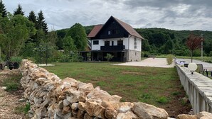 Une maison à deux étages, avec un mur de pierre au premier plan et une colline boisée en arrière-plan.