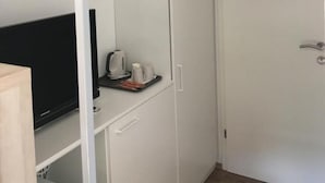 A compact room with a white wardrobe, a small table with a kettle and cups, and a television on a shelf.