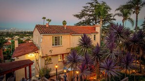 A two-story house with a red-tiled roof, surrounded by lush landscaping including palm trees and agave plants.