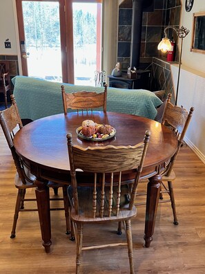 A round wooden dining table with six chairs, a decorative centerpiece, and a view of a forest through a glass door.