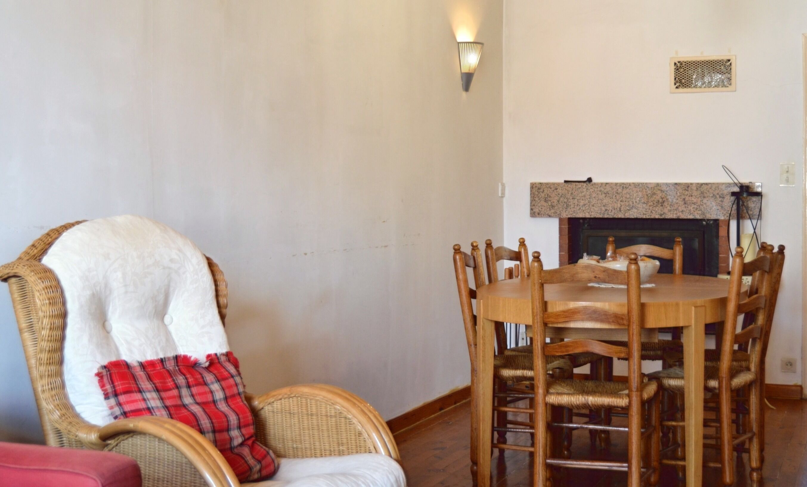A dining area with a wooden table and chairs, a wicker chair with a plaid blanket, and a fireplace.