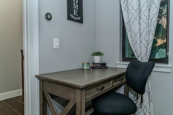 A room with a desk, chair, a potted plant, a thermostat, and a window with a curtain.