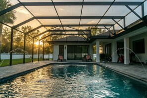 A swimming pool with a covered patio area, chairs, and a view of the sunset.