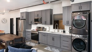 A modern kitchen with grey cabinets, stainless steel appliances, and a dining area with a wooden table and blue cushions. - Industrial Retreat Where Recreation Abounds! Dogs OK, Pool Table, BBQ & More! (Bozeman)