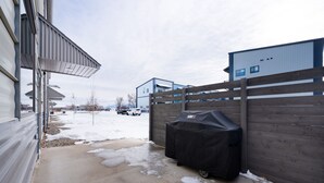 A covered outdoor area with a grill, a wooden fence, and a building in the background. - Industrial Retreat Where Recreation Abounds! Dogs OK, Pool Table, BBQ & More! (Bozeman)