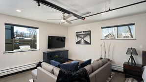 A modern living room with a large sofa, a flat-screen TV, and a ceiling fan. - Industrial Retreat Where Recreation Abounds! Dogs OK, Pool Table, BBQ & More! (Bozeman)