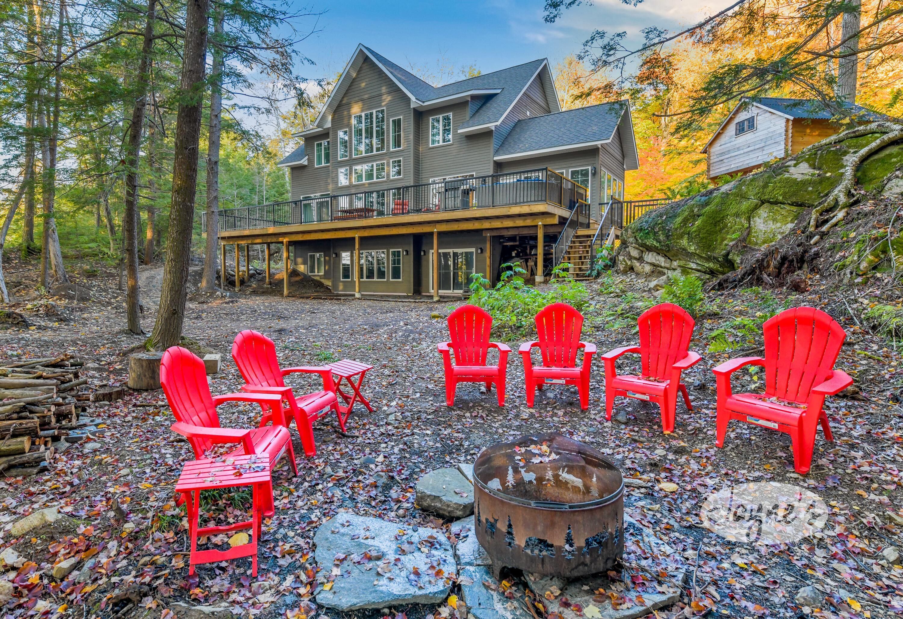 A large house with a deck, surrounded by trees and a fire pit with red chairs.