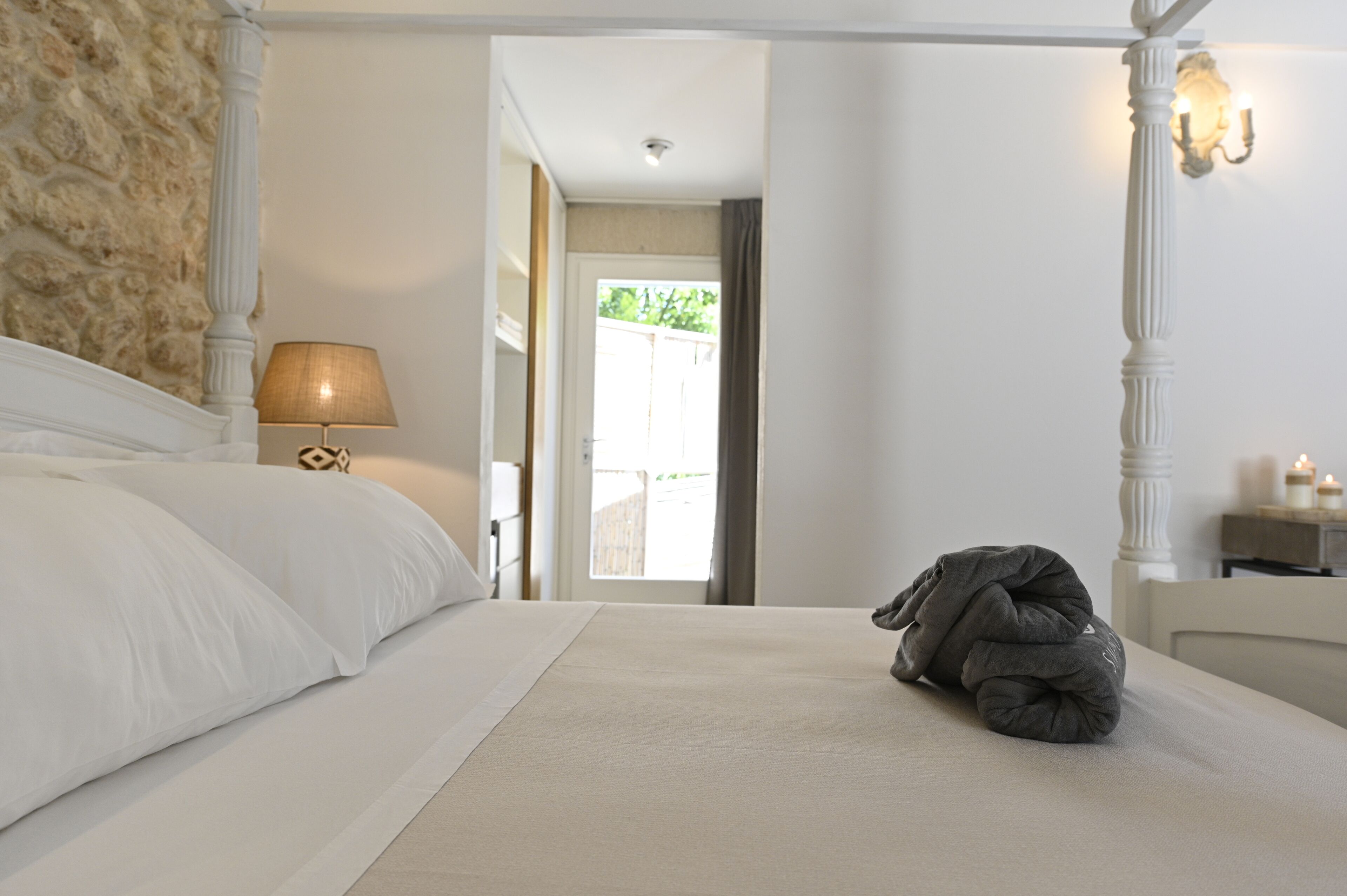 Un dormitorio con cama con dosel, lámpara de noche, mesita de noche con velas y vistas al exterior a través de una puerta.