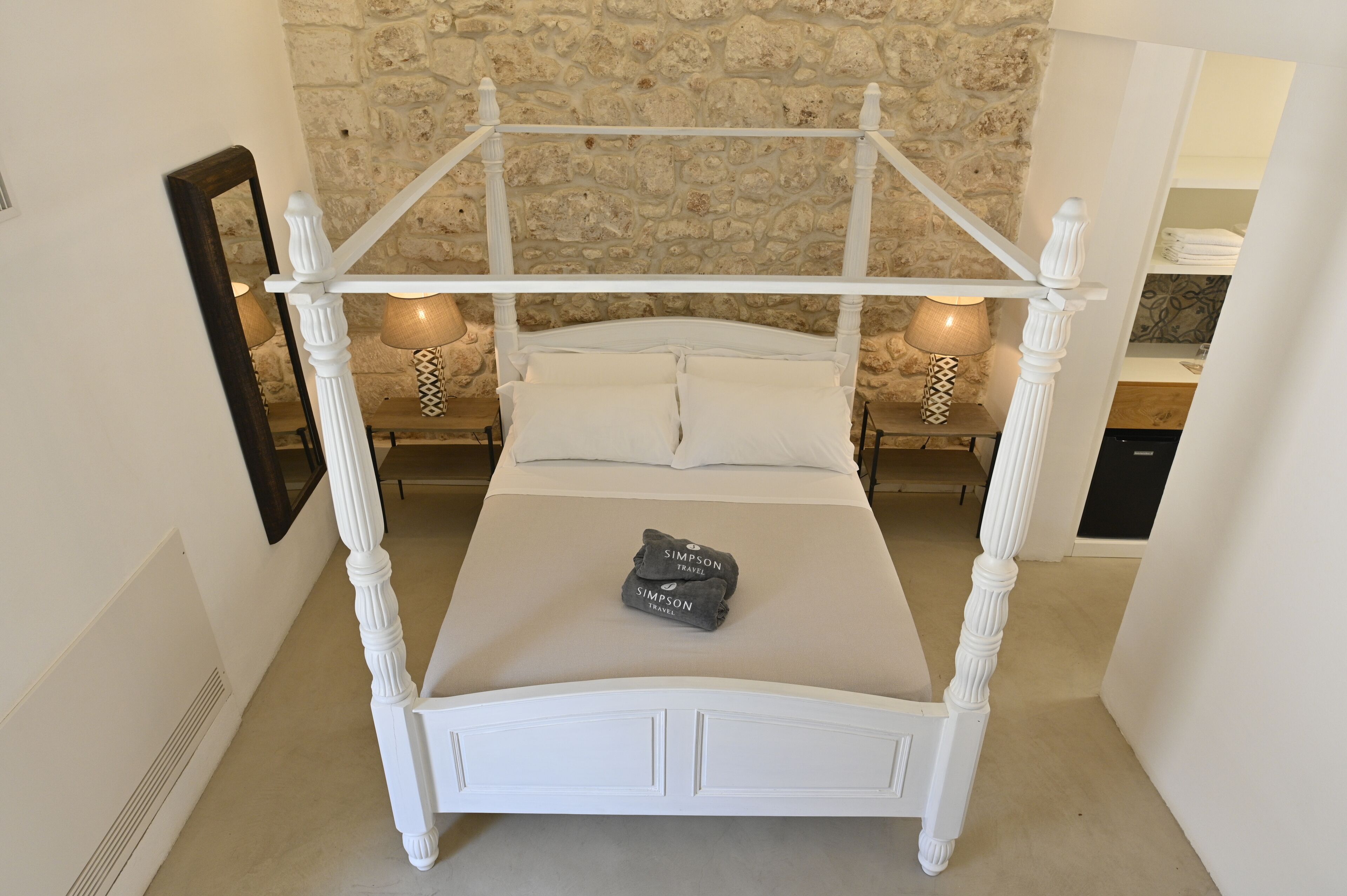 A white four-poster bed with a stone wall background, a mirror on the left, and two bedside lamps.