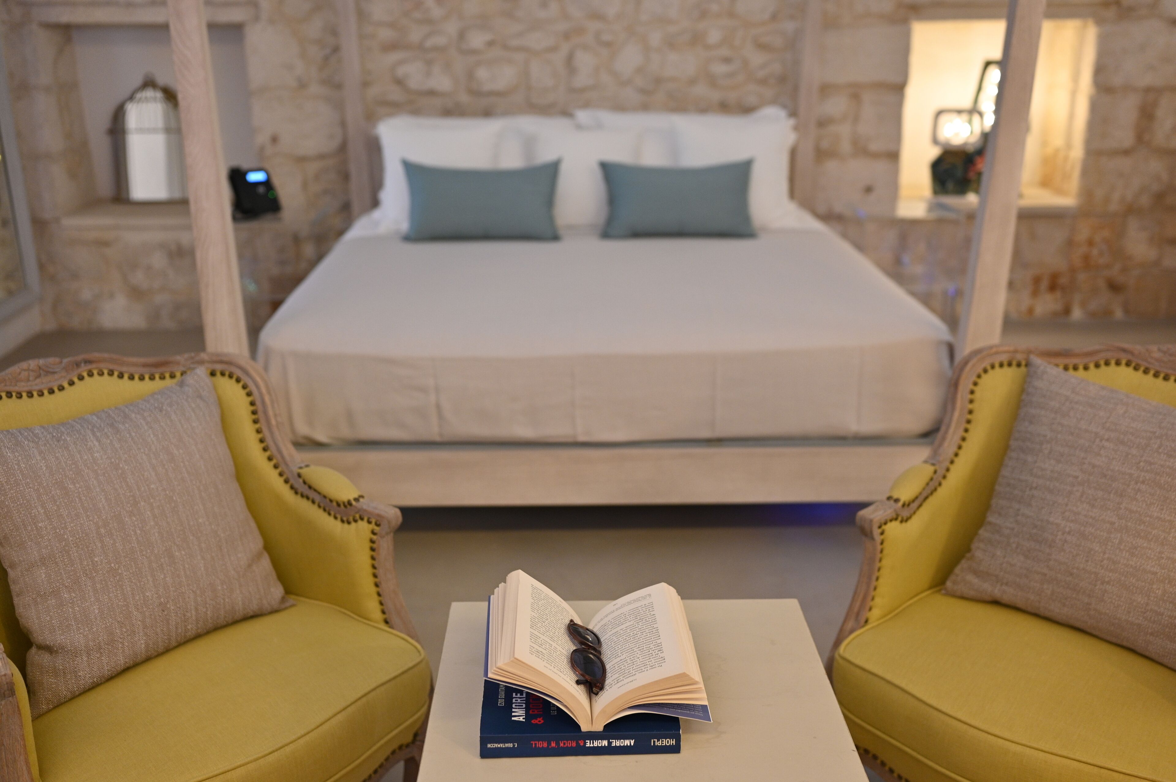 A bedroom with a bed, two armchairs, a bedside table with a book, and a stone wall.
