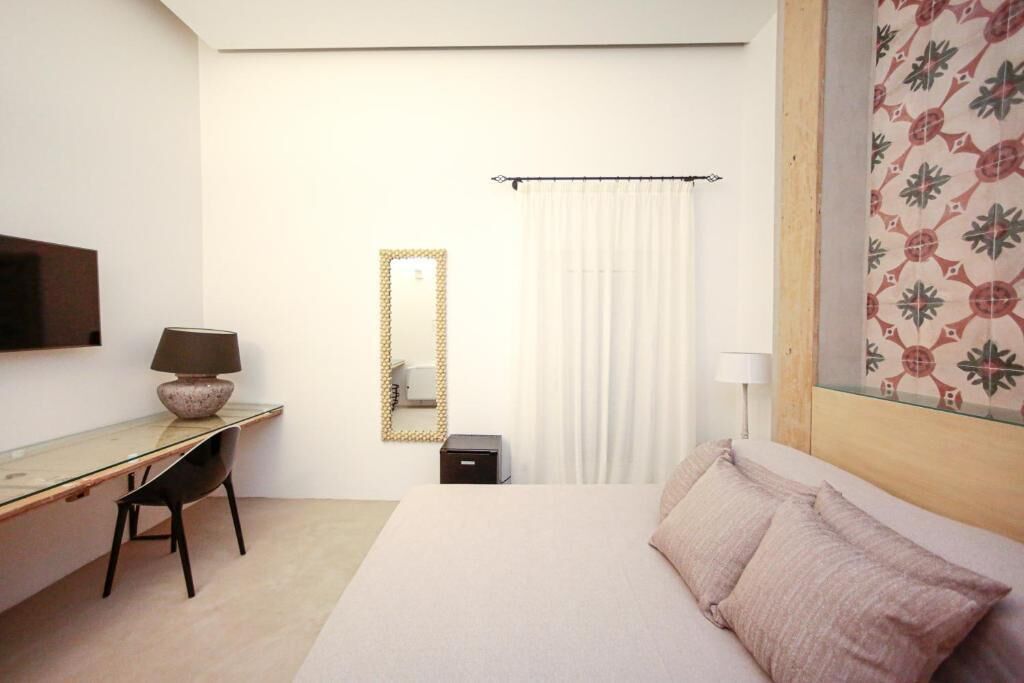 A bedroom with a bed, a desk, a chair, a TV, a mirror, and a window with curtains.