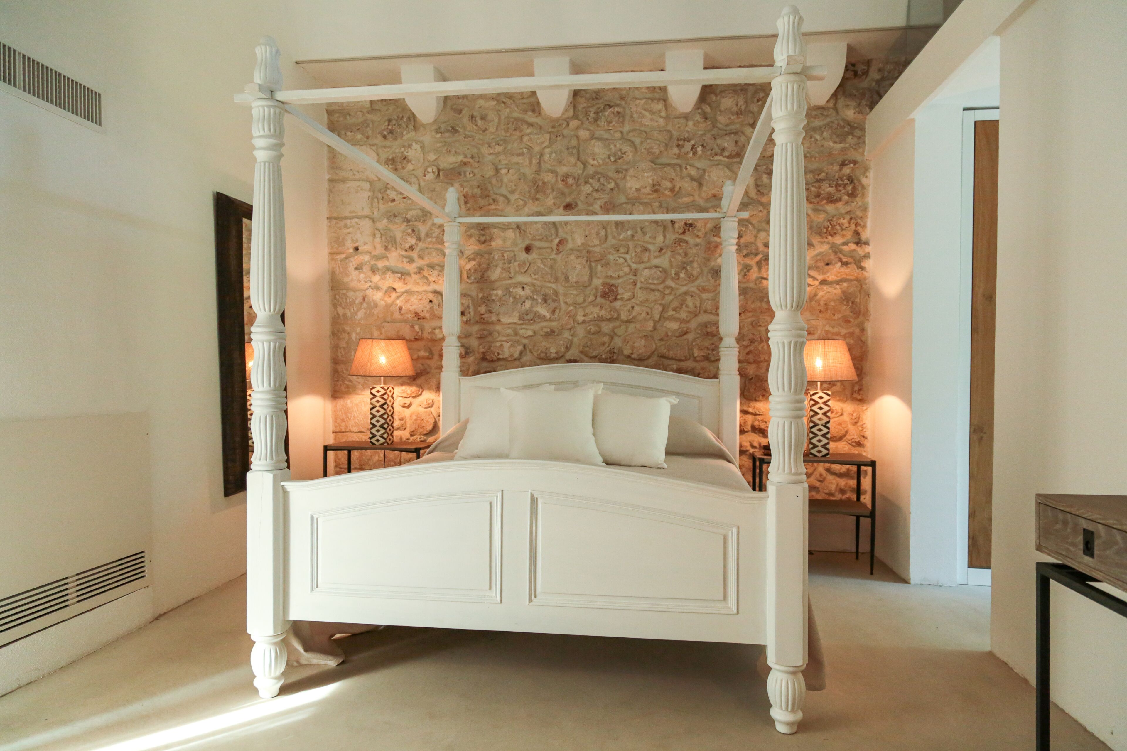 Una cama con dosel frente a una pared de piedra, dos lámparas de noche y una mesita de madera.