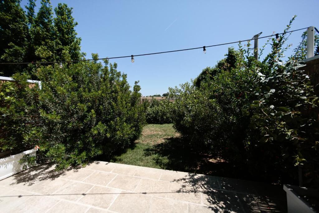 Un patio empedrado con luces colgantes, rodeado de follaje abundante y un cielo despejado.