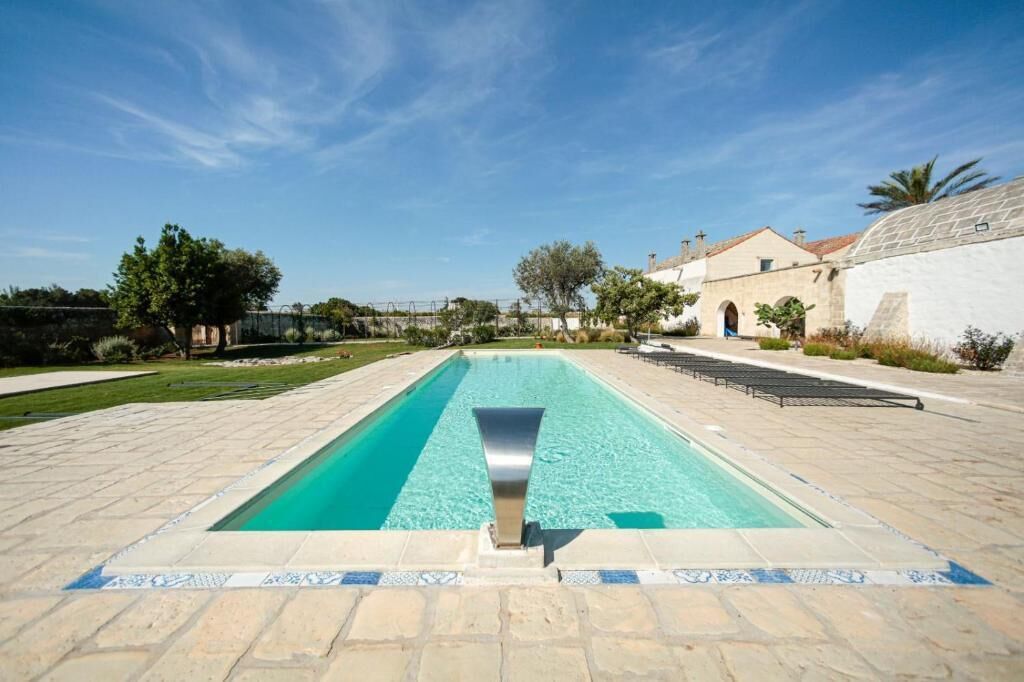 A rectangular pool with a clear blue water feature, surrounded by a paved area with stone tiles and a few trees.