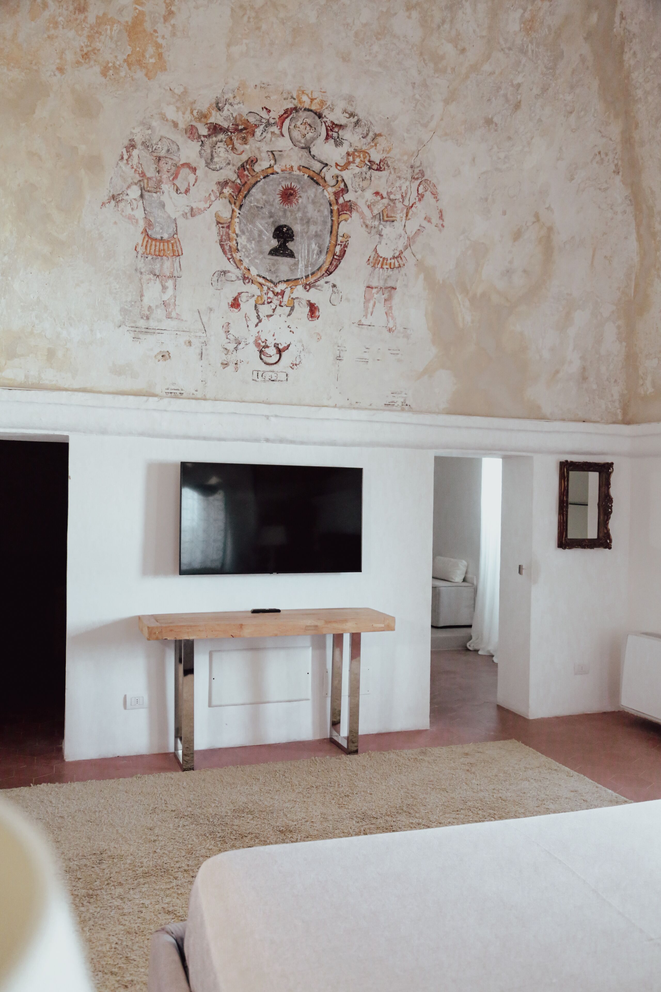Een slaapkamer met een aan de muur bevestigde televisie, een houten bankje, een bed en een muur met een decoratief fresco.
