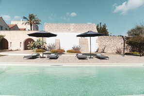 Zona de piscina con sillas de descanso y sombrillas, un muro de piedra y un edificio con arcos al fondo.