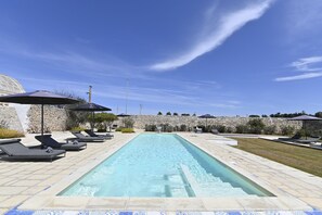 Piscina con sillas de descanso y sombrillas, rodeada de muros de piedra y un cielo despejado.