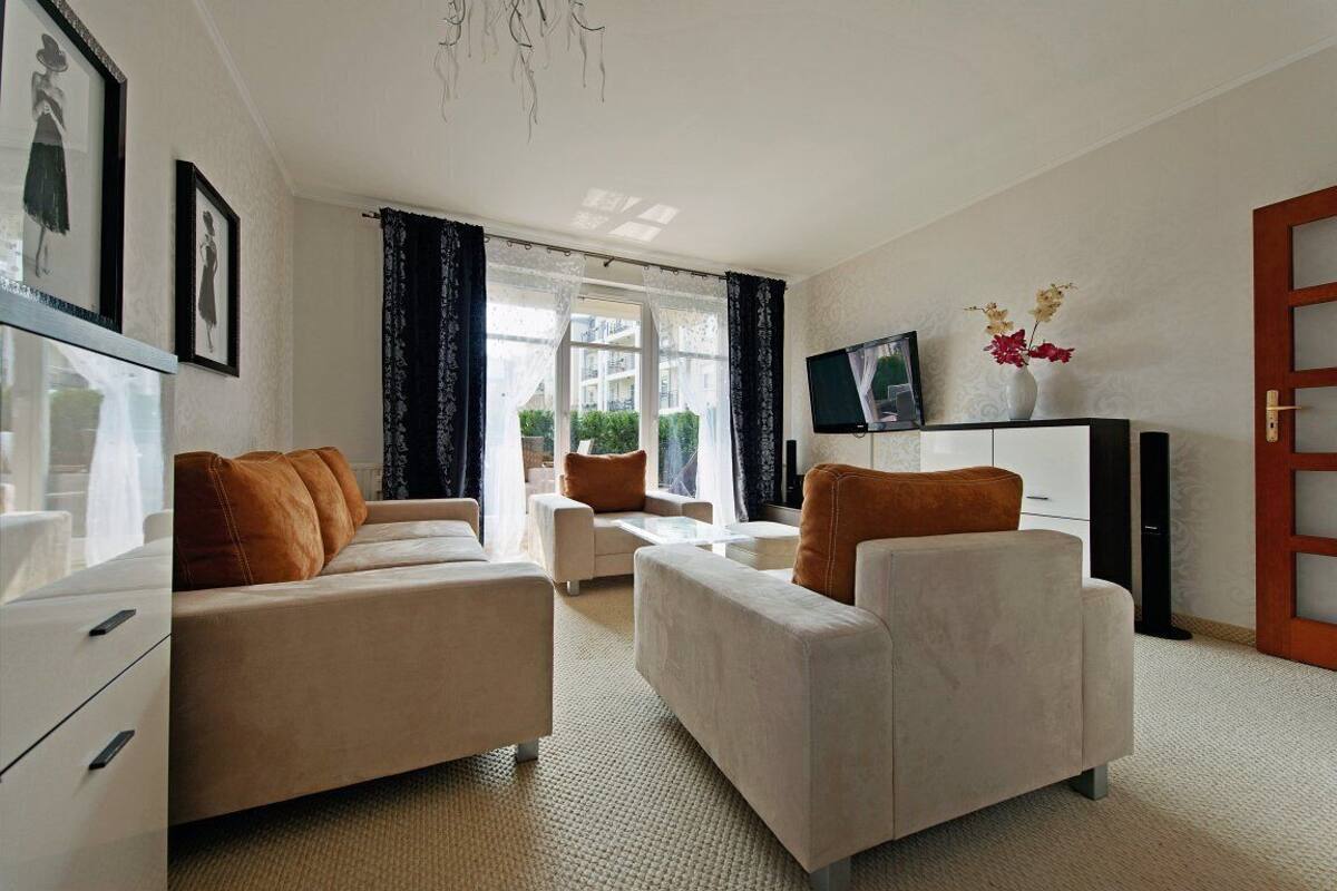 A modern living room with a sofa, armchairs, a coffee table, and a TV. There is a window with curtains, a door, and a plant on the windowsill.
