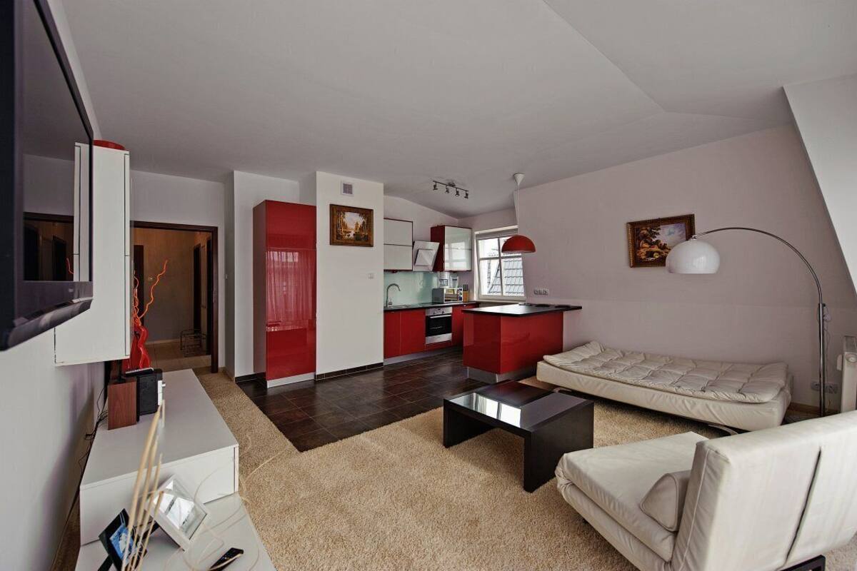 A modern living room with a sofa, a coffee table, and a TV. There is a kitchen area with red cabinets and a dining table.