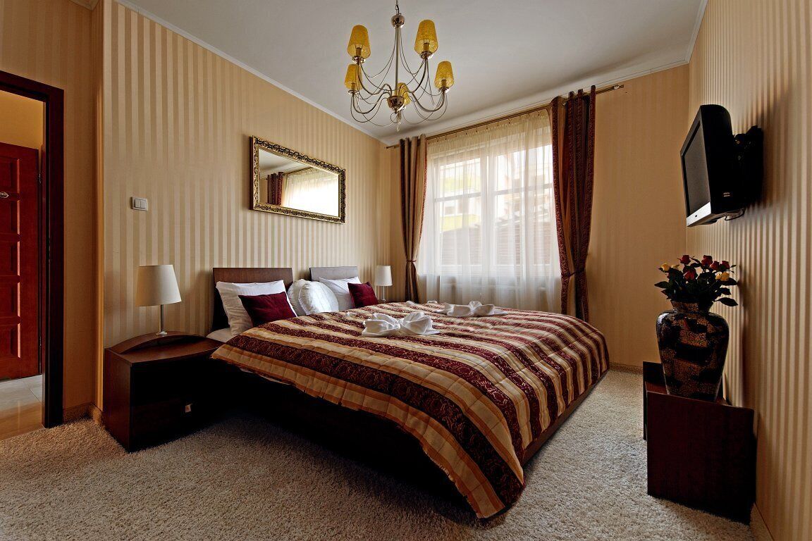 A hotel room with a large bed, a TV mounted on the wall, a chandelier, and a vase with flowers on a side table.