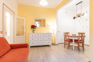 Een kamer met een bank, een witte dressoir, een eettafel met stoelen, een spiegel en een wandklok.