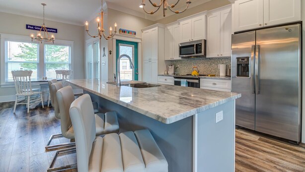 A modern kitchen with white cabinets, a stainless steel refrigerator, a marble island with bar stools, and a chandelier. - Bright beach house steps from the sand & memories (Long Beach)