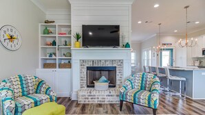 A living room with a fireplace, two patterned armchairs, a yellow ottoman, and a TV on a white mantel. - Bright beach house steps from the sand & memories (Long Beach)