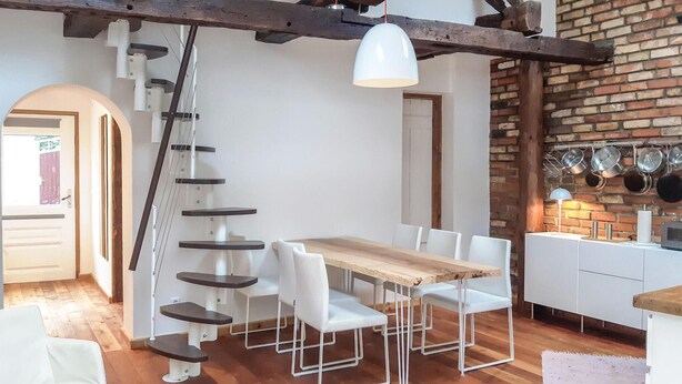 Een eetruimte met een houten tafel en witte stoelen, een witte hanglamp en een bakstenen muur met aanbehangende potten.