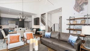 A modern living room with a leather sofa, a dining area with a kitchen in the background, and a staircase leading to another level. - Ski-in/out from Back Door, Mtn Modern Moonlight Basin "Wildwood Luxury Chalet", Hot Tub, Garage (Big Sky)