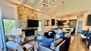 A modern living room with a stone fireplace, a flat-screen TV, and a dining area with a kitchen in the background. - Ski-in/out from Back Door, Mtn Modern Moonlight Basin "Wildwood Luxury Chalet", Hot Tub, Garage (Big Sky)