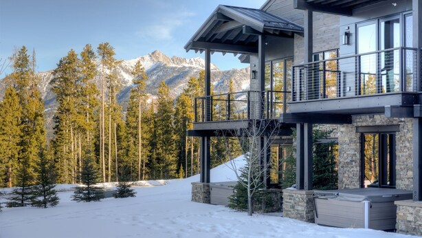 A modern two-story house with a stone facade, large windows, and a balcony overlooking a snowy mountain landscape. - Ski-in/out from Back Door, Mtn Modern Moonlight Basin "Wildwood Luxury Chalet", Hot Tub, Garage (Big Sky)