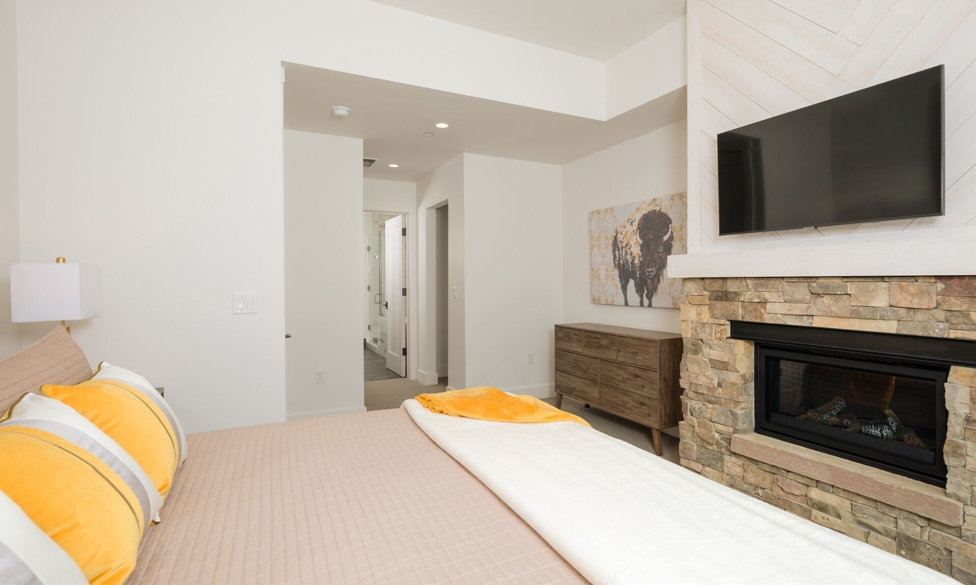 A bedroom with a bed, a TV mounted on the wall, a fireplace, and a door leading to another room.
