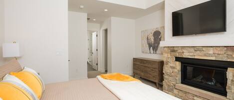 A bedroom with a bed, a TV mounted on the wall, a fireplace, and a door leading to another room.