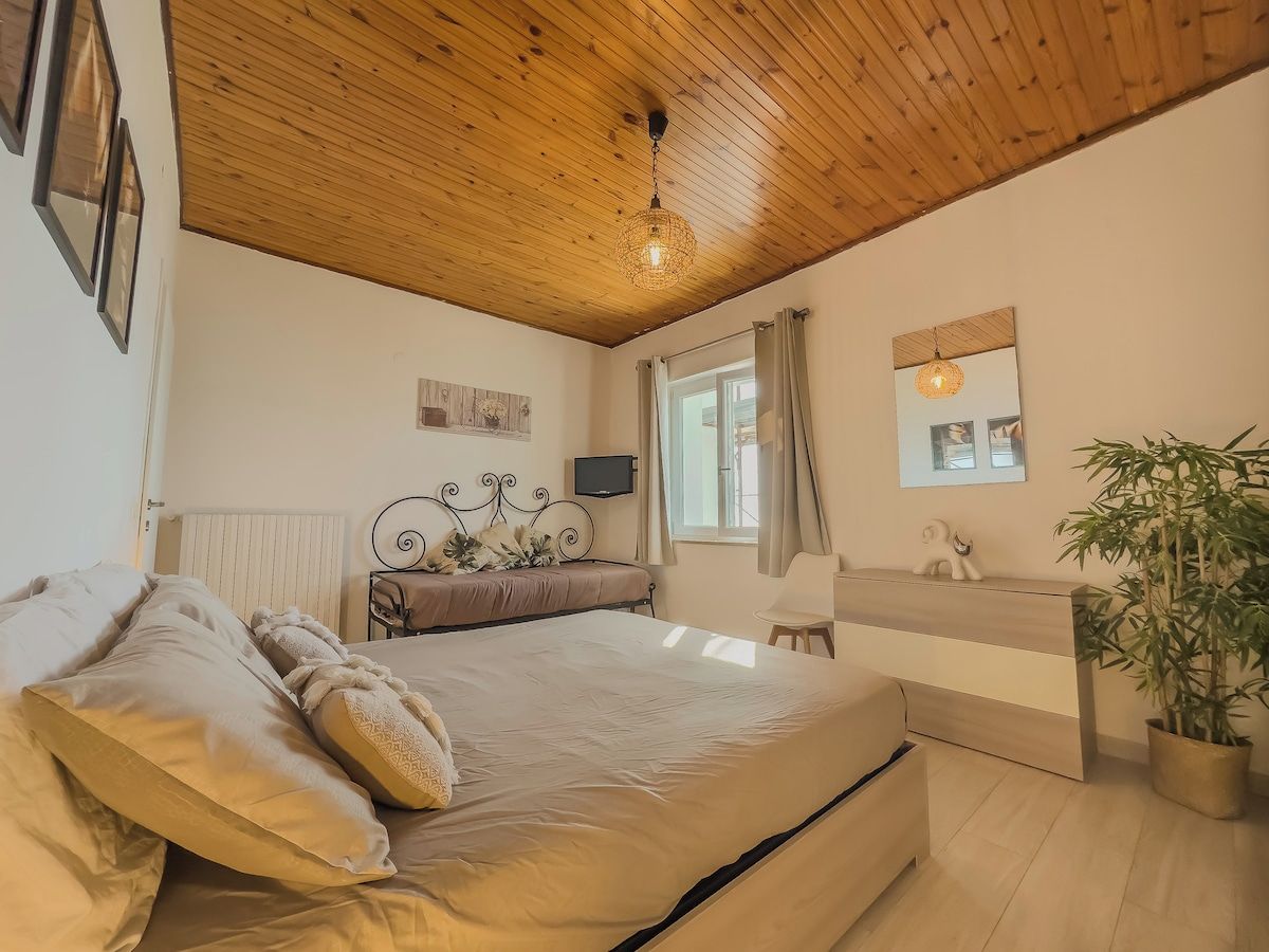 A bedroom with a wooden ceiling, a bed with pillows, a bedside table, a mirror, a window with curtains, and a potted plant.