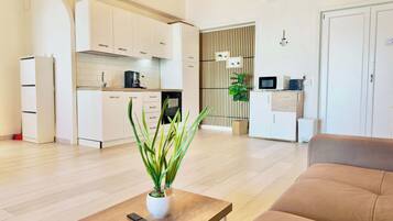 A modern living room with a sofa and a coffee table, a kitchen with white cabinets, and a potted plant.
