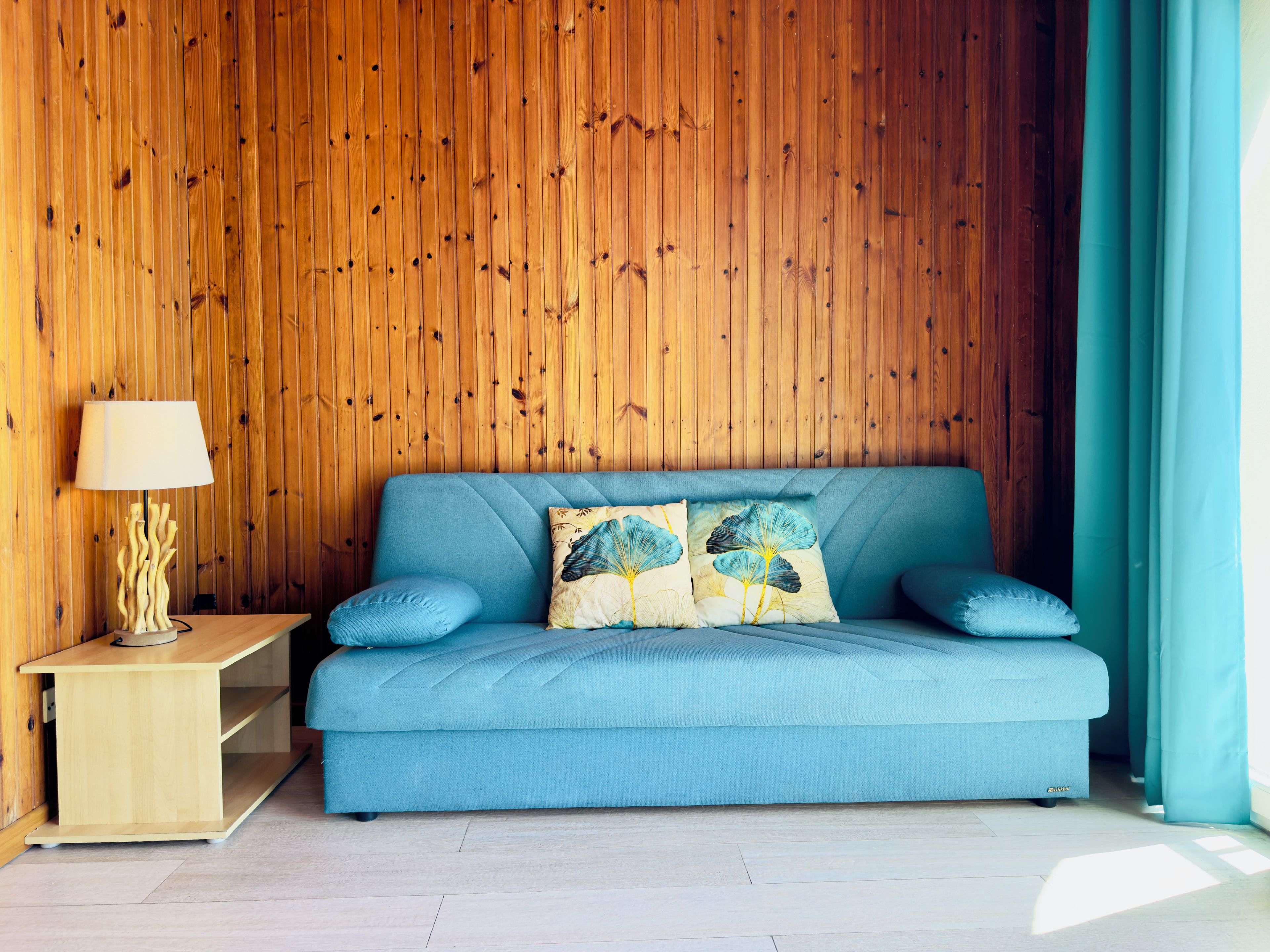 Ein Zimmer mit einem blauen Sofa, einer Holzwand, einer Lampe und einem Vorhang.